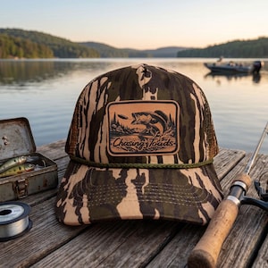 May include: A camouflage baseball cap with a leather patch featuring a fish jumping out of the water, with the text "Chasing Toads." The hat has a mesh back and a green rope detail. Fishing gear and a lake are in the background.