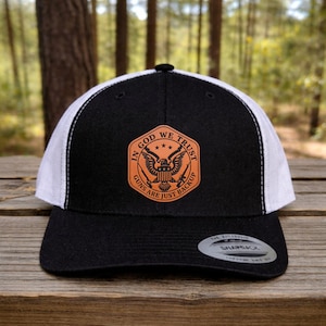 May include: Black and white trucker hat with a black brim and a white mesh back. The front features a brown patch with the text "IN GOD WE TRUST" and "GUNS ARE JUST BACKUP" surrounding an eagle and crossed rifles.