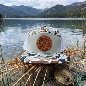 May include: A camouflage-patterned trucker hat with a white mesh back and a light green front panel. The hat features a circular leather patch with the text "Fish or a Buzz... I'm Catching Something" and a fish graphic.
