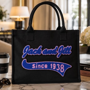 May include: Black tote bag with the text "Jack and Jill" in blue script with pink trim, and "Since 1938" on a blue banner. The bag has two black handles and is sitting on a marble surface.