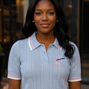 May include: A light blue short-sleeved polo shirt featuring a vertical cable knit texture, a collar with white and pink stripes, and matching striped sleeve cuffs. The chest displays an embroidered logo with pink and dark blue horizontal lines.