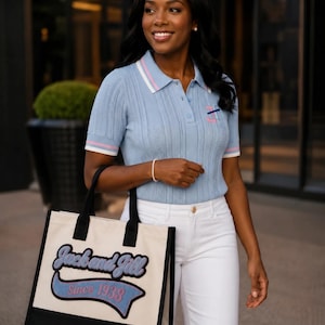 May include: A light blue short-sleeved polo shirt with white and pink striped collar and sleeve cuffs, paired with white pants and a cream-colored canvas tote bag featuring black trim and the text Jack and Jill Since 1938 in blue and pink.