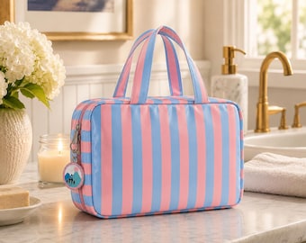 May include: A rectangular travel bag with vertical stripes in light blue and pink. The bag has two handles and a zipper closure. A small round charm with a mountain scene hangs from the zipper.