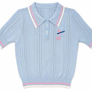 May include: A light blue short-sleeved polo shirt with a cable knit texture. The collar and sleeve cuffs feature white and pink stripes. A small embroidered logo in pink and dark blue is positioned on the left chest area below the collar.