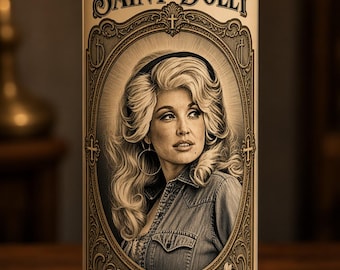 May include: A tall cylindrical glass prayer candle filled with white wax. The label features a sepia-toned portrait of Dolly Parton, the text "SAINT DOLLY," and the quote "If you see someone without a smile, give 'em yours" with dates.