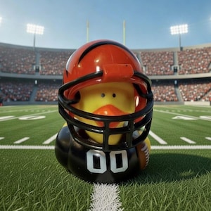 May include: A yellow rubber duck wearing a football helmet with black face mask and orange stripes. The duck is on a green football field with white yard lines, in front of a stadium filled with spectators.