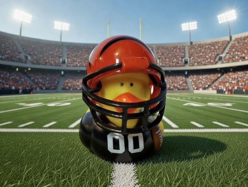 May include: A yellow rubber duck wearing a football helmet with black face mask and orange stripes. The duck is on a green football field with white yard lines, in front of a stadium filled with spectators.