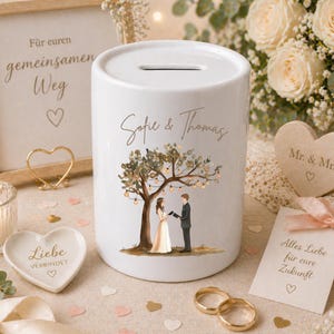 May include: A white ceramic money box featuring a cylindrical shape with a coin slot on top. The front displays a printed illustration of a couple standing under a tree with hanging lights, with the names Sofie & Thomas written above.
