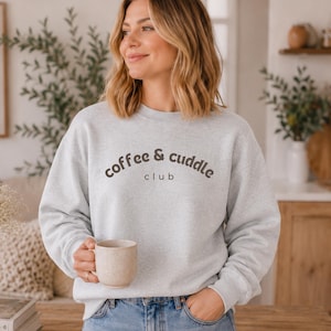 May include: A light gray crewneck sweatshirt featuring the dark brown text coffee & cuddle club printed across the chest. The garment has long sleeves with ribbed cuffs and a ribbed neckline, providing a standard fit for casual wear.