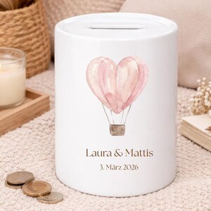May include: White ceramic money bank with a heart-shaped hot air balloon design in shades of pink. The bank has a slot at the top and features the names "Laura & Mattis" and the date "3. M&auml;rz 2026" printed on the front.