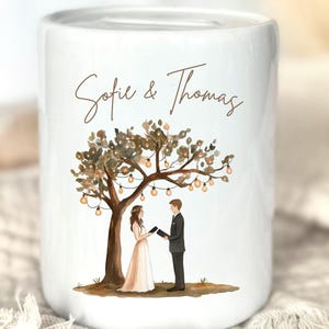 May include: A white ceramic money box featuring a printed illustration of a couple standing under a tree decorated with string lights. The text "Sofie & Thomas" is printed in brown script above the couple, who are holding small black books.