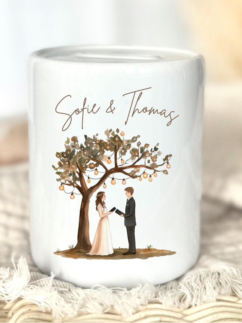 May include: A white ceramic money box featuring a printed illustration of a couple standing under a tree decorated with string lights. The text "Sofie & Thomas" is printed in brown script above the couple, who are holding small black books.