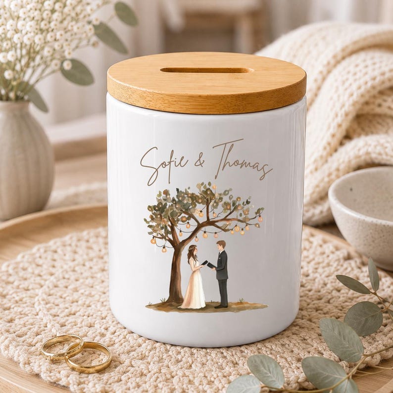 May include: A white ceramic money box with a cylindrical shape and a light brown wooden lid featuring a coin slot. The front displays a printed illustration of a couple standing under a tree with hanging lights and the text Sofie & Thomas.
