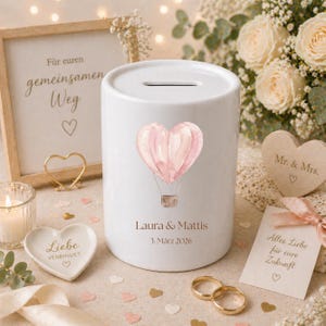 May include: A white ceramic money bank with a slot on top, decorated with a pink heart-shaped hot air balloon. The bank is personalized with names and a date. Wedding-themed decor includes a framed sign, rings, and a heart-shaped dish.