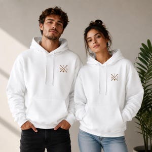 May include: A white hooded sweatshirt featuring a brown graphic on the left chest. The graphic consists of two crossed arrows with the letters T, W, and S positioned between the arrows and a small heart icon at the bottom center of the design.