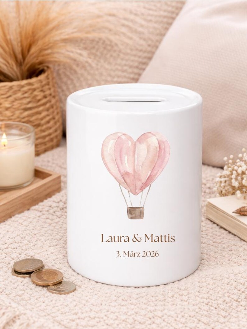 May include: White ceramic money bank with a heart-shaped hot air balloon design in shades of pink. The bank has a slot at the top and features the names "Laura & Mattis" and the date "3. M&auml;rz 2026" printed on the front.