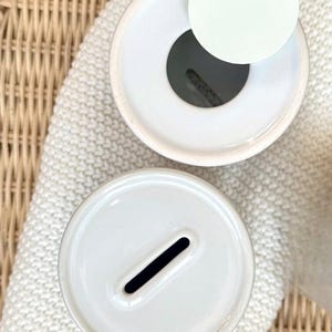 May include: Two white ceramic piggy banks viewed from directly above. One bank features a circular opening with a separate white lid placed beside it, while the other bank has a narrow, rectangular coin slot centered on its smooth, flat top surface.