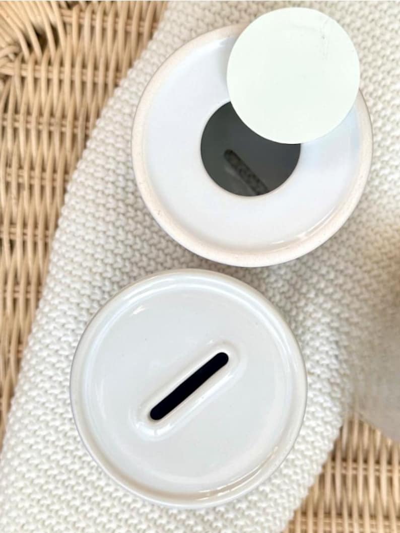 May include: Two white ceramic piggy banks viewed from directly above. One bank features a circular opening with a separate white lid placed beside it, while the other bank has a narrow, rectangular coin slot centered on its smooth, flat top surface.