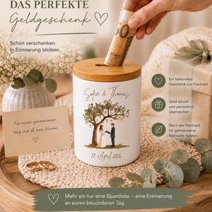 May include: A white ceramic money box with a wooden lid featuring a printed illustration of a couple under a tree with string lights, the names Sofie & Thomas, and the date 27. April 2026. The container is cylindrical with a coin slot on top.