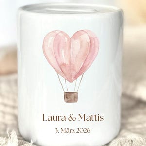 May include: White ceramic bank with a watercolor illustration of a pink heart-shaped hot air balloon. The bank has the names "Laura & Mattis" and the date "3. M&auml;rz 2026" printed in brown.