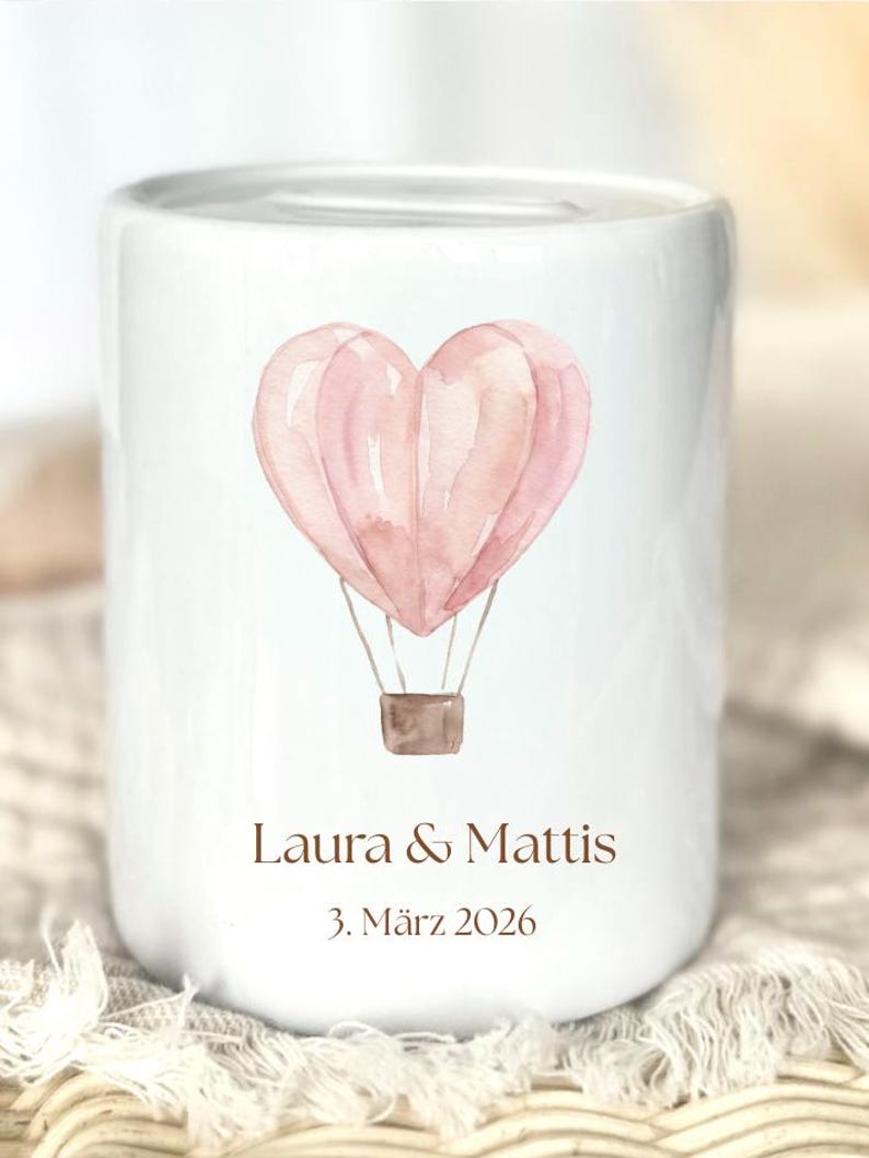 May include: White ceramic bank with a watercolor illustration of a pink heart-shaped hot air balloon. The bank has the names "Laura & Mattis" and the date "3. M&auml;rz 2026" printed in brown.