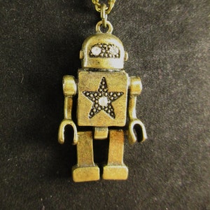 May include: A gold robot pendant necklace with a star on the chest and rhinestone accents. The robot is wearing a chain necklace.