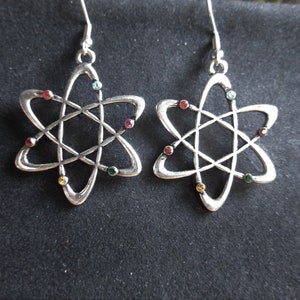 Może przedstawiać: Para srebrnych kolczyków w kształcie atomu z kolorowymi kryształkami. Kolczyki mają kształt stylizowanego atomu z centralnym okręgiem i pięcioma punktami.