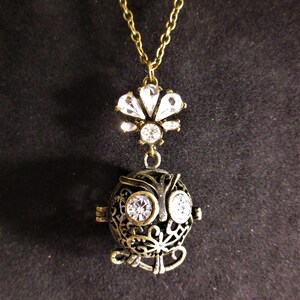 Op de afbeelding: Een gouden ketting met een kristallen uil bedel. De uil heeft heldere strass steentjes als ogen en een kristallen bloemenontwerp op het lichaam. De ketting heeft een kristallen bloemenhanger boven de uil.