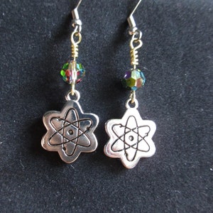 Może przedstawiać: Srebrne kolczyki w kształcie atomu z iryzującymi zielonymi i fioletowymi koralikami. Symbol atomu to stylizowana reprezentacja atomu z jądrem i krążącymi elektronami.