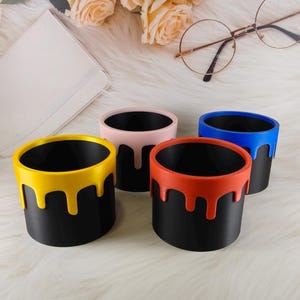 May include: Four black cylindrical containers with colorful paint drip designs in yellow, pink, red, and blue. The containers are arranged on a white, fluffy surface, with a bouquet of peach roses and glasses in the background.