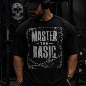 Pode incluir: Camiseta preta com a frase "MASTER THE BASIC" em letras brancas desgastadas, emoldurada por um desenho de corrente. A pessoa usa boné e fones de ouvido pretos. O fundo mostra equipamentos de ginástica.