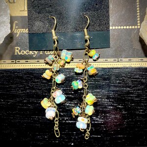 May include: Dangling earrings with gold-colored hooks and chains. Each earring features clusters of colorful, iridescent opal stones in shades of yellow, blue, green, and white. The earrings are displayed against a dark background.