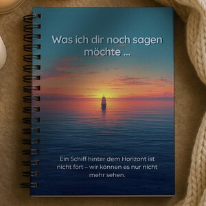 Könnte beinhalten: Ein Spiralnotizbuch mit einer Sonnenuntergangsszene und einem Segelboot auf dem Meer. Der Einband hat deutschen Text. Mehrere brennende Kerzen und eine cremefarbene Strickdecke befinden sich im Hintergrund.