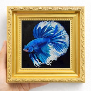 May include: A framed painting of a blue and white betta fish against a black background. The fish has flowing fins and is set in a gold ornate frame. The artwork is held by a hand.