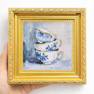 May include: An oil painting in a gold ornate frame depicting a stack of three white teacups with blue floral patterns. The cups feature gold rims and are rendered with visible brushstrokes against a light gray and blue textured background.
