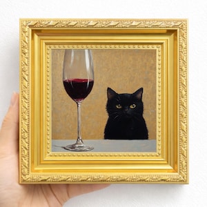 Op de afbeelding: Een vierkant ingelijst schilderij met een zwarte kat naast een hoog wijnglas gevuld met rode wijn. Het kunstwerk zit in een sierlijke, getextureerde gouden lijst met een herhalend geometrisch patroon langs de buitenrand.