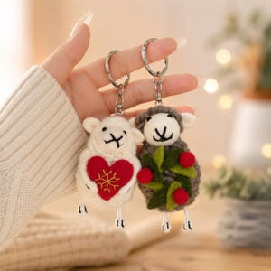 May include: Two felted sheep keychains with metal rings. One sheep is white and holds a red heart with a gold embroidered snowflake. The other sheep is gray and holds a green wreath with red berries. Both have embroidered faces and dangling legs.