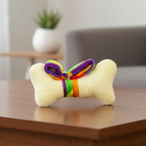 Puede incluir: Un juguete de hueso para perros de color amarillo claro, con una cinta de arco&iacute;ris atada alrededor del centro. La cinta presenta tonos de morado, verde, naranja y amarillo. El juguete est&aacute; dise&ntilde;ado para mascotas.