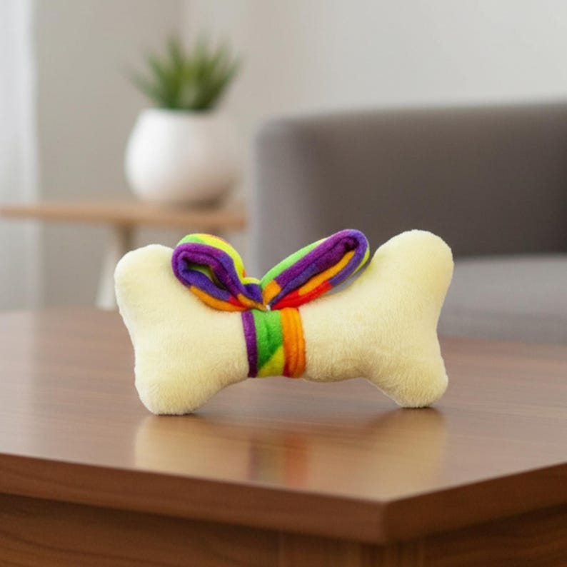 Puede incluir: Un juguete de hueso para perros de color amarillo claro, con una cinta de arco&iacute;ris atada alrededor del centro. La cinta presenta tonos de morado, verde, naranja y amarillo. El juguete est&aacute; dise&ntilde;ado para mascotas.