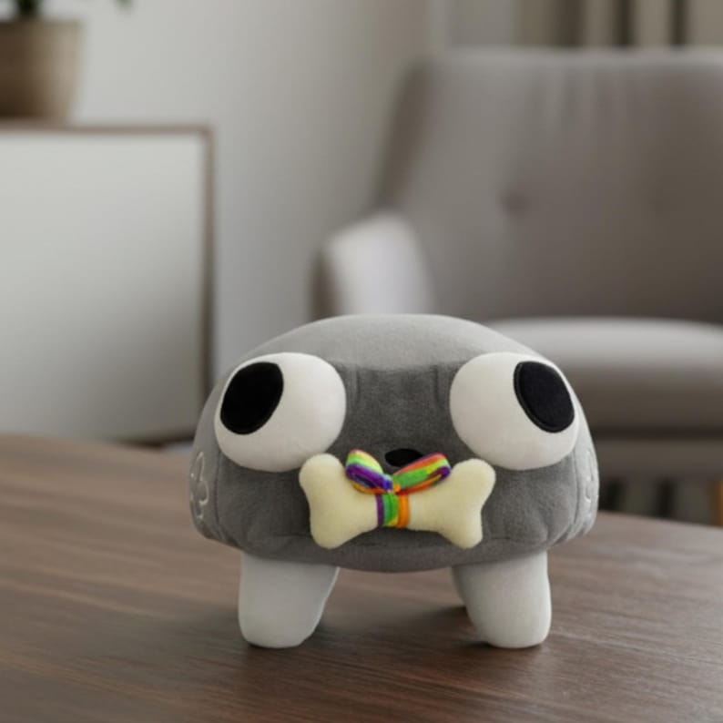 Puede incluir: Un peluche gris con forma de roca, con grandes ojos blancos y pupilas negras. Tiene un hueso color crema con una cinta arco&iacute;ris. El juguete tiene pies blancos y est&aacute; sobre una superficie de madera.