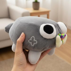 Puede incluir: Un peluche gris con ojos grandes y caricaturescos y un accesorio colorido en forma de mariposa. El juguete tiene un dise&ntilde;o de tr&eacute;bol de cuatro hojas en un lado y peque&ntilde;os pies blancos. El juguete est&aacute; en la mano de una persona.