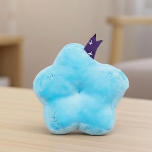 Pode incluir: Um brinquedo de pel&uacute;cia em forma de estrela azul claro com um detalhe roxo e branco. O brinquedo macio tem costuras vis&iacute;veis e uma pequena pe&ccedil;a decorativa roxa com cruzes brancas. O brinquedo est&aacute; em uma superf&iacute;cie de madeira.