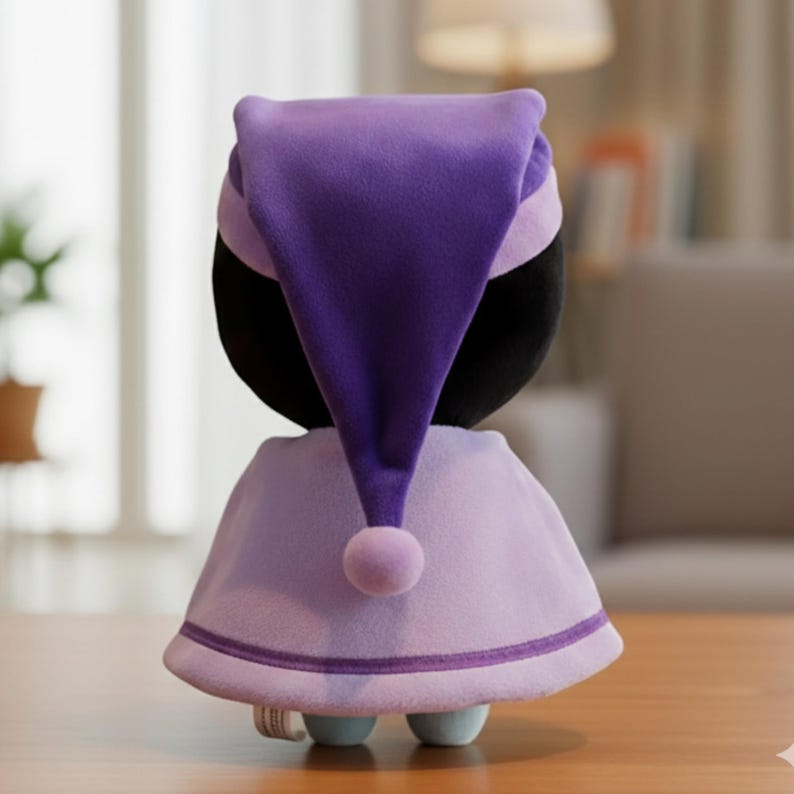 Pode incluir: Uma boneca de pel&uacute;cia com cabe&ccedil;a preta e gorro roxo. A boneca usa uma capa roxa clara com acabamento roxo escuro e um pompom combinando. A boneca est&aacute; em uma superf&iacute;cie de madeira.