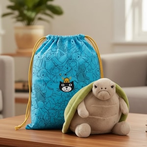 Peut inclure: Une peluche en forme de tortue beige avec une carapace verte est posée à côté d'un sac bleu à cordon. Le sac présente un motif de personnages de dessins animés et un motif de chat noir. Le sac a des cordons jaunes. La tortue a une expression renfrognée.
