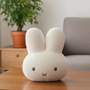 以下が含まれることがあります： 2つの直立した耳を持つ、オフホワイトのウサギ型のぬいぐるみクッション。ウサギは黒い点の目と、口を表す黒い刺繍の「x」があります。クッションは木製のテーブルの上にあり、背景には植物とソファがあります。