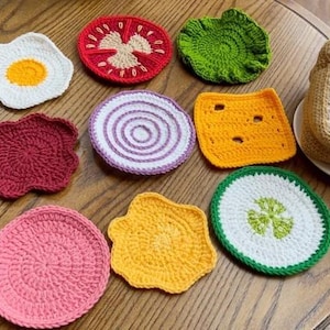 May include: A collection of handmade crochet food-themed coasters, including a fried egg, tomato slice, lettuce, red beet, onion, cheese, cucumber slice, and a stack of toast. The coasters are arranged on a wooden table.