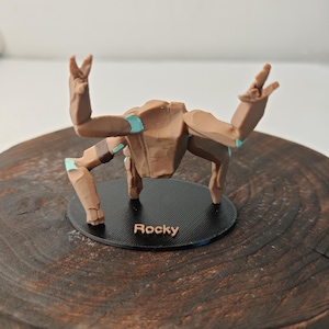 May include: A brown, articulated figurine with raised arms and legs, mounted on a black circular base. The figurine has light blue accents and the name "Rocky" in gold lettering on the base. The figurine is displayed on a dark wooden surface.