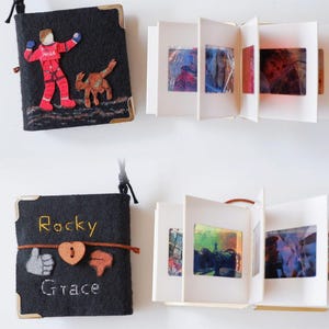 Könnte beinhalten: Zwei kleine, handgefertigte Bücher aus schwarzem Filz mit Eckenschützern und angebrachten Kordeln. Ein Buch zeigt einen Astronauten und einen Hunde-Applikation, das andere hat die Namen "Rocky" und "Grace" mit einem Herz und Daumen hoch/runter-Symbolen bestickt. Die Bücher haben farbenfrohe Innenseiten.