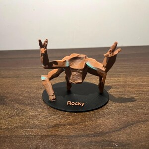 May include: A brown, stylized figurine of a creature with outstretched arms and legs, mounted on a black circular base. The base has the name "Rocky" printed on it. The figurine has blue accents.