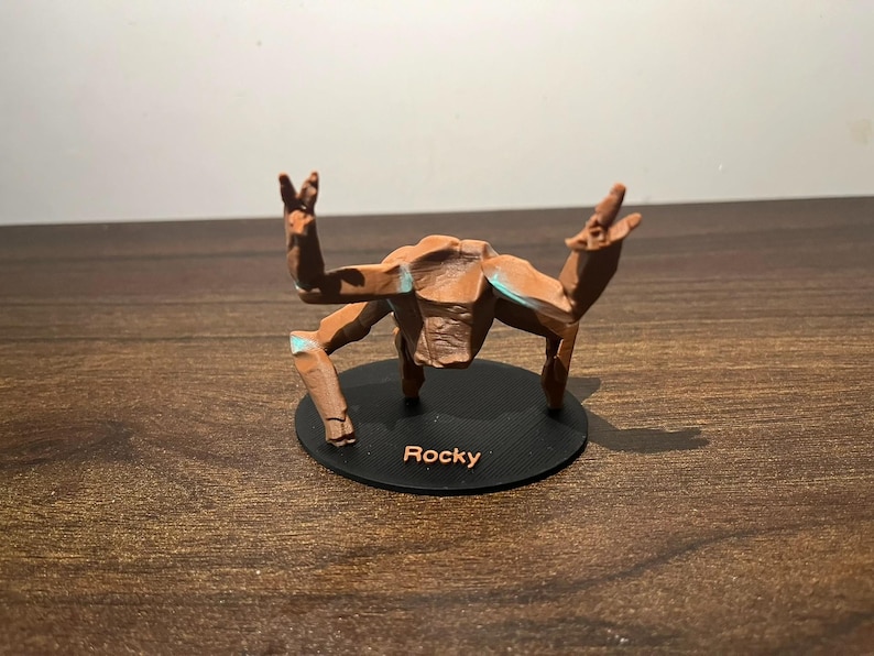 May include: A brown, stylized figurine of a creature with outstretched arms and legs, mounted on a black circular base. The base has the name "Rocky" printed on it. The figurine has blue accents.